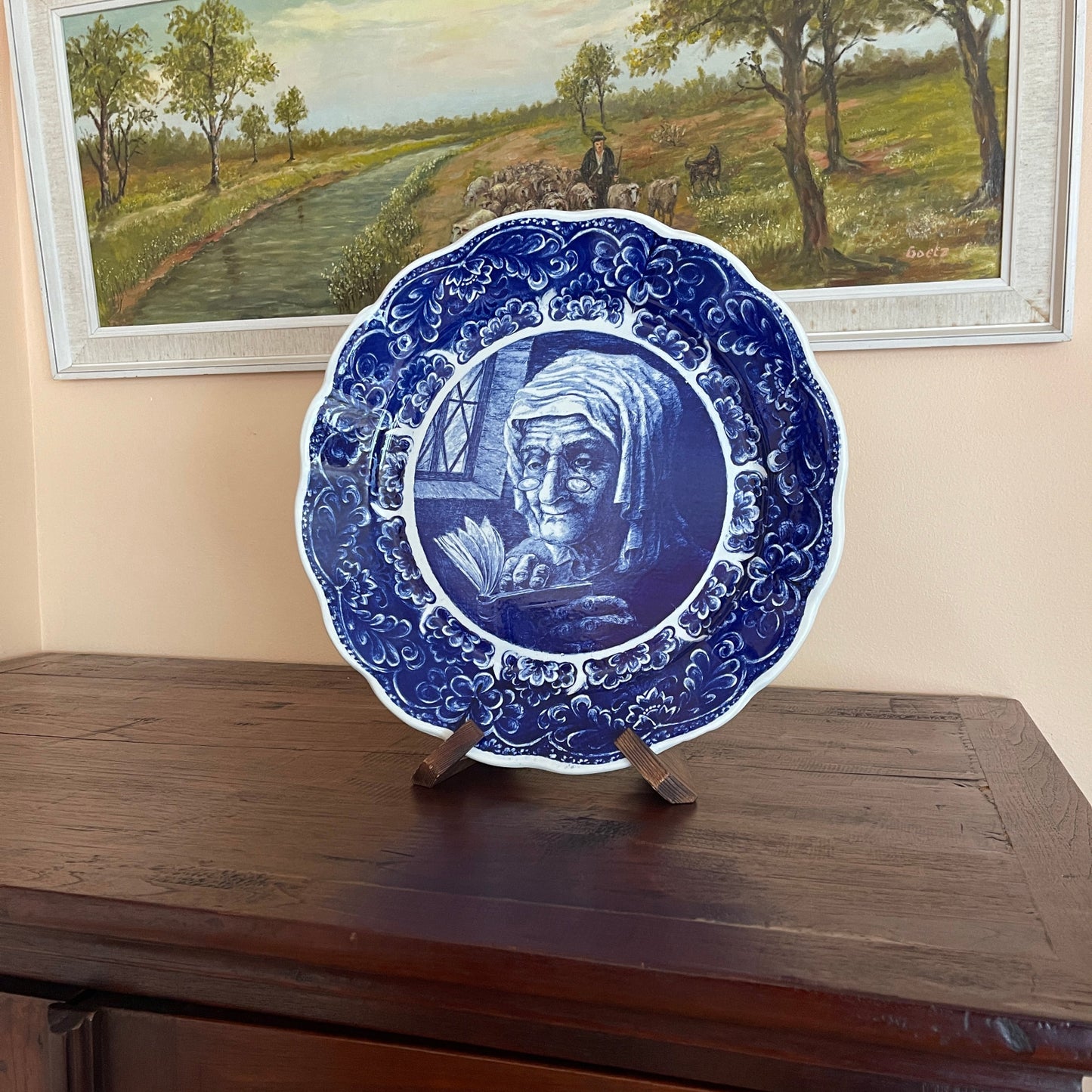 Blue decorative plate with a portrait on a wooden stand, framed landscape painting in the background.