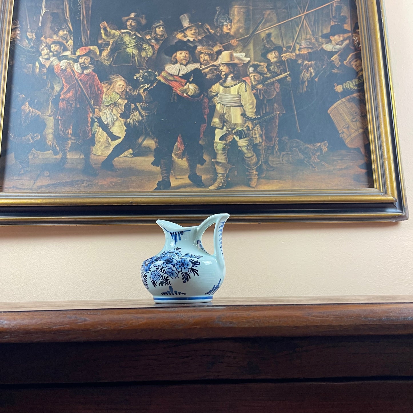 Small blue and white ceramic pitcher on a wooden surface with a large painting in the background.