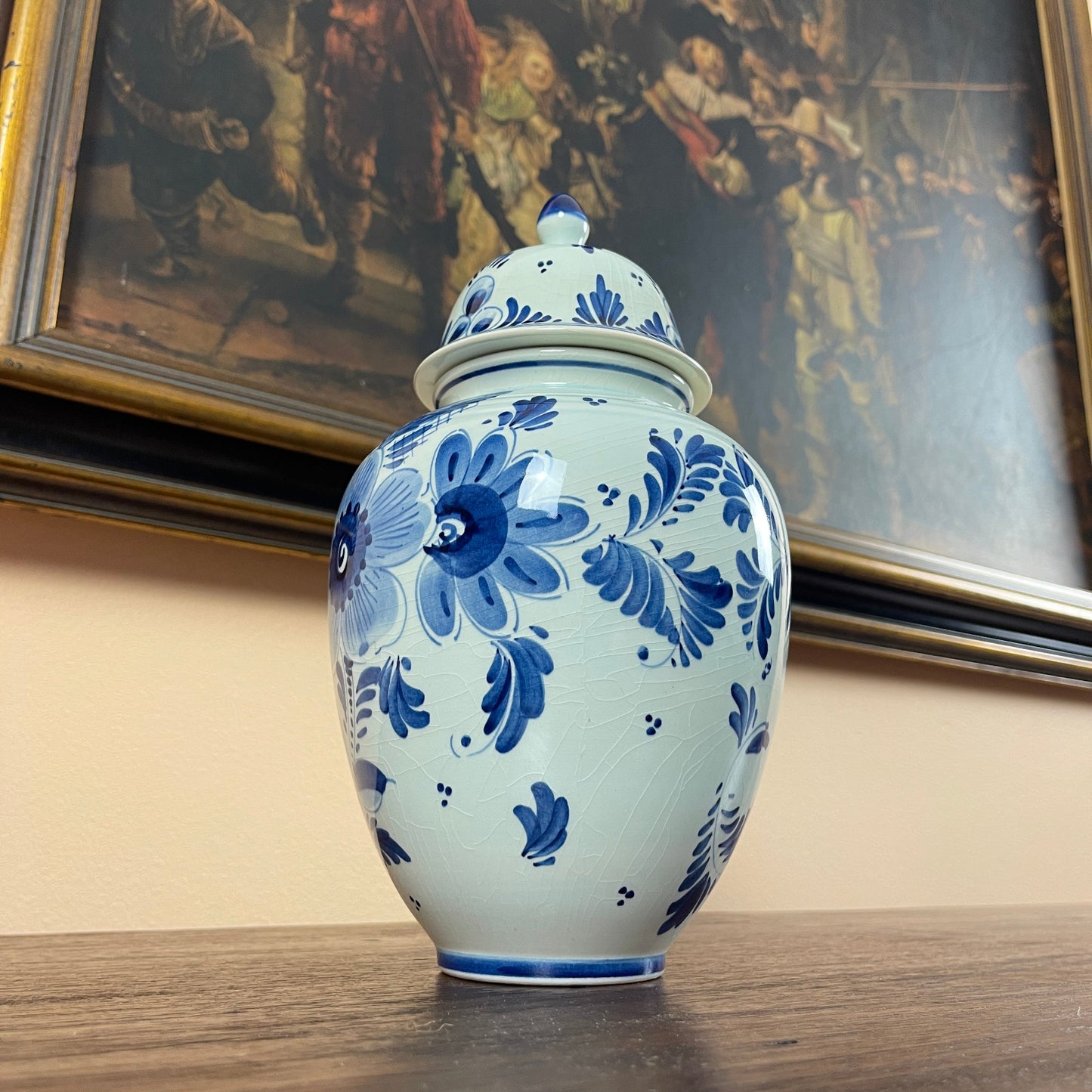 Blue and white floral-patterned ceramic jar on a wooden surface with a painting in the background
