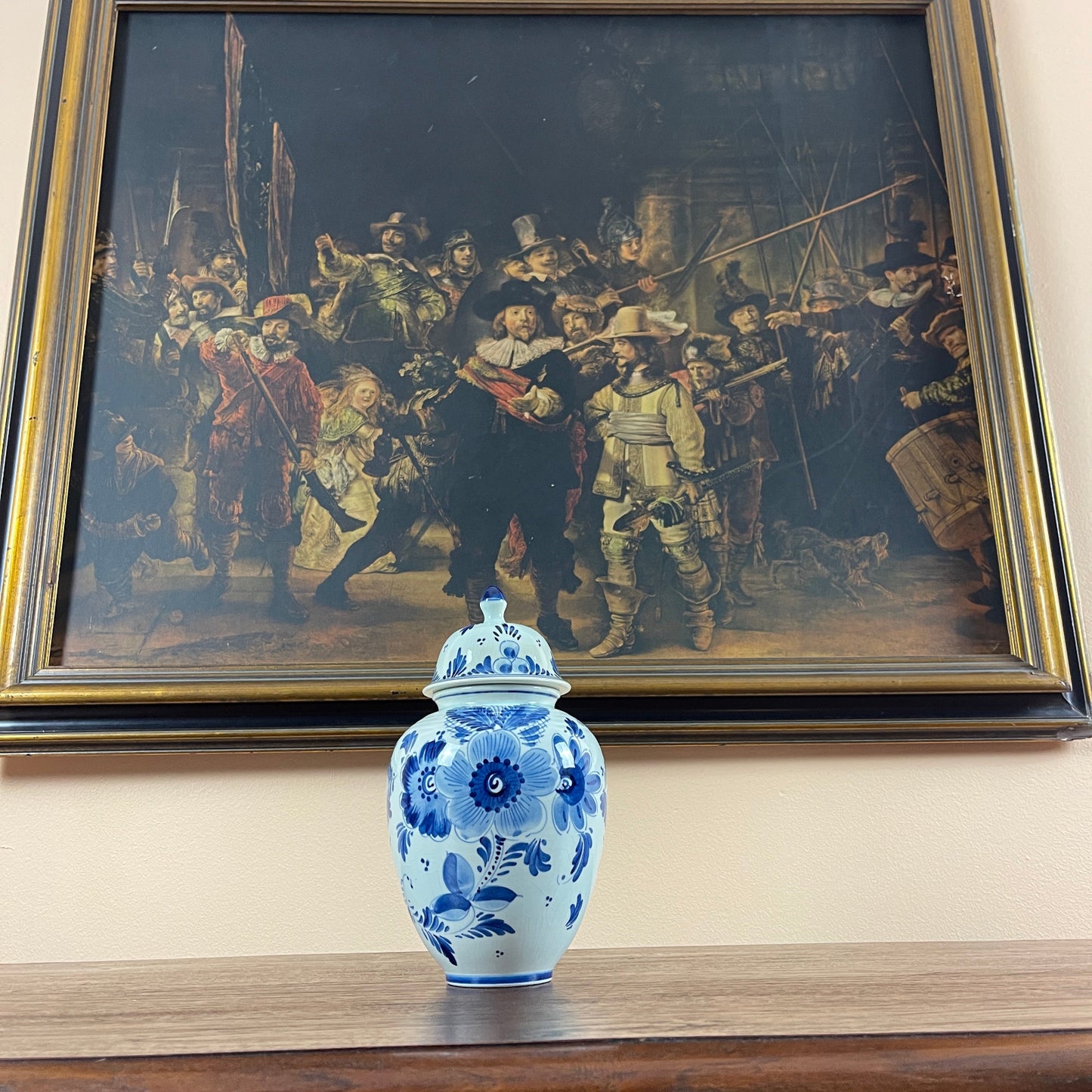 Blue and white floral-patterned vase on a surface with a framed painting of 'The Night Watch' in the background.