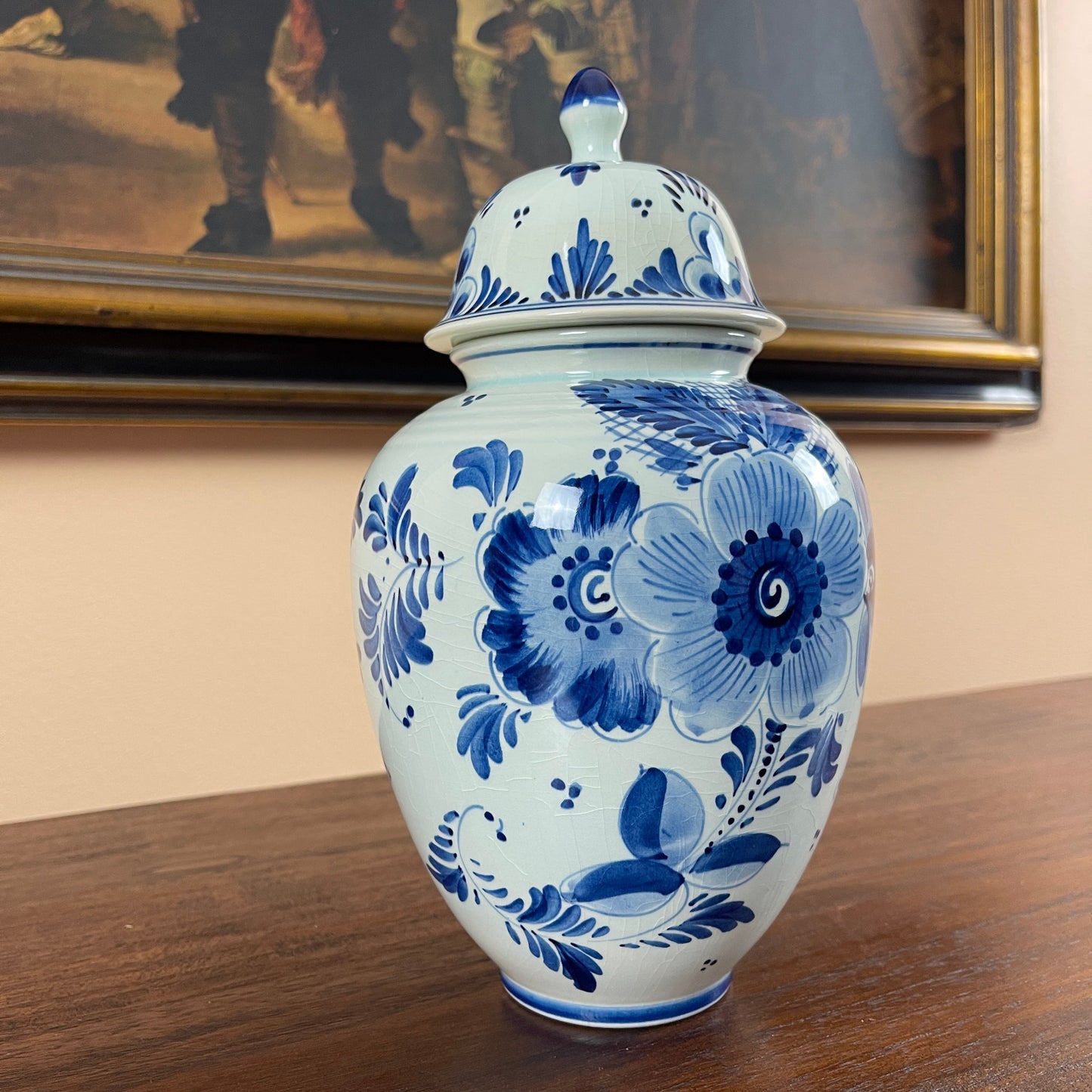 Blue and white floral-patterned ceramic jar on a wooden surface with a painting in the background.