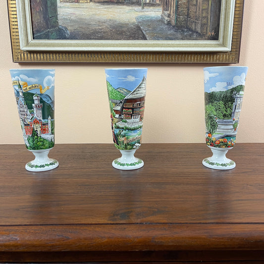 Three scenic-themed glasses on a wooden surface with a framed painting in the background.