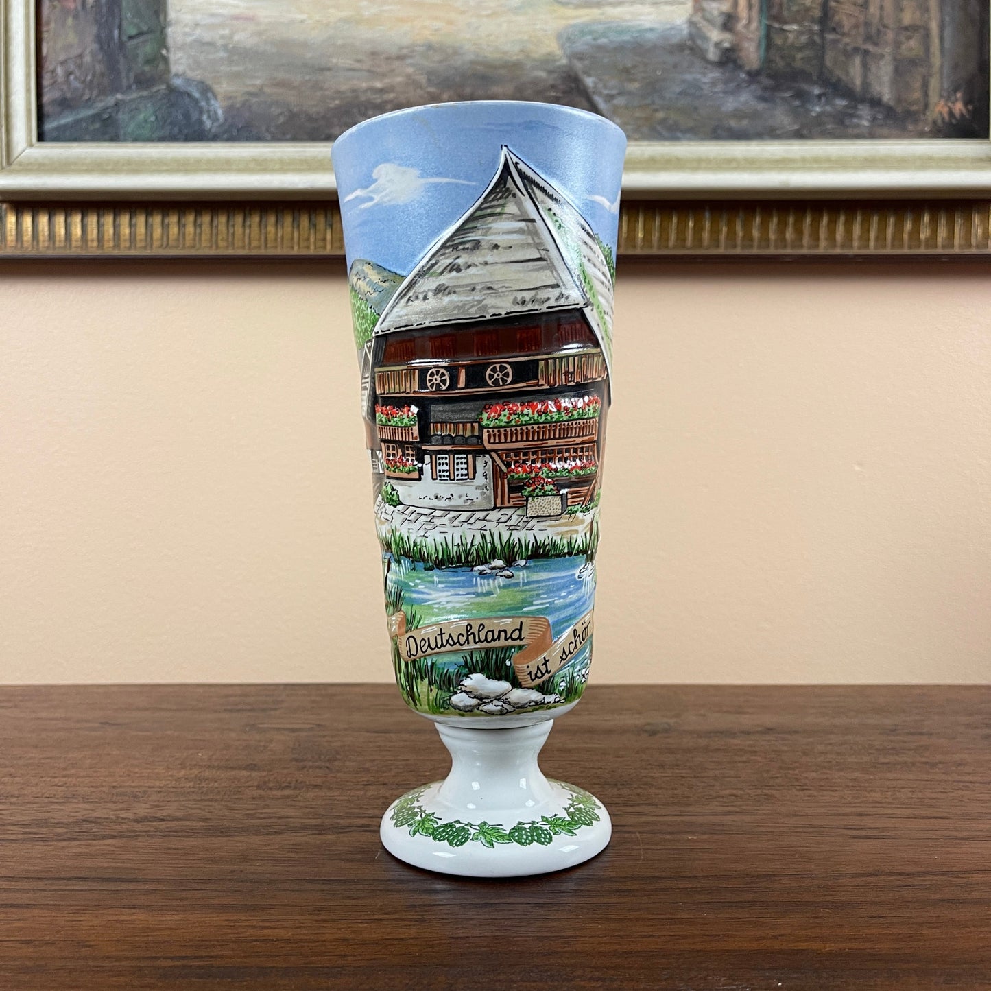 Decorative glass with a painted scene of a house and landscape on a wooden surface.