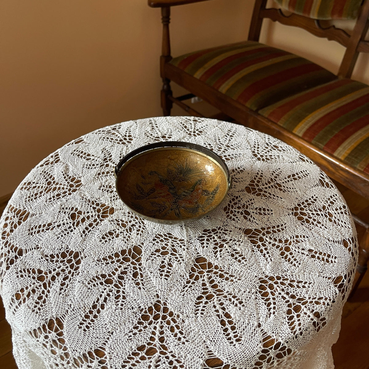 Round table with lace tablecloth and brass bowl, wooden chair in the background
