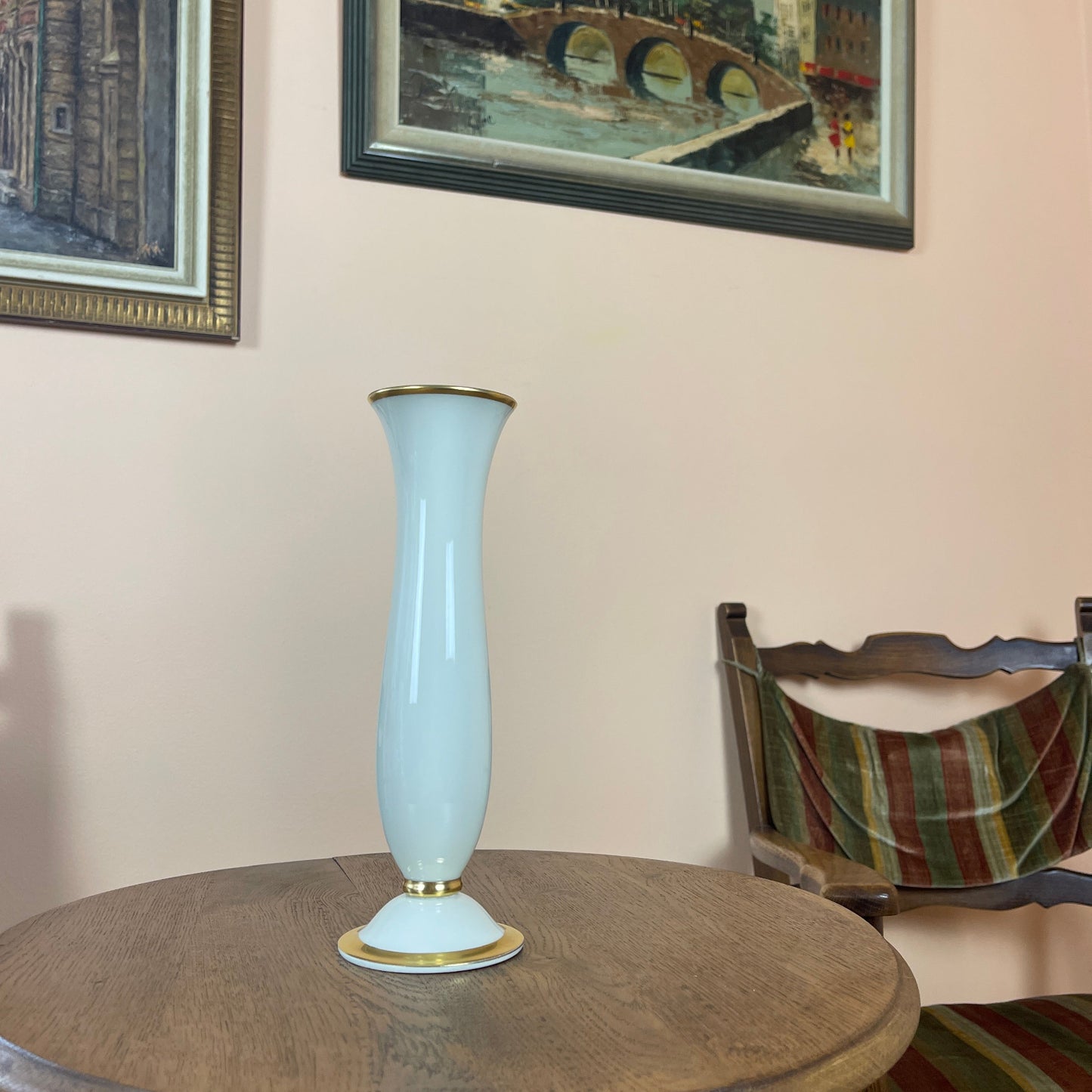 Light blue vase with gold accents on a round wooden table, with a chair and framed picture in the background.