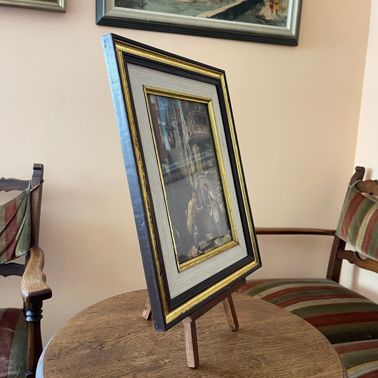 Framed picture on a stand with chairs and a wall with framed art in the background