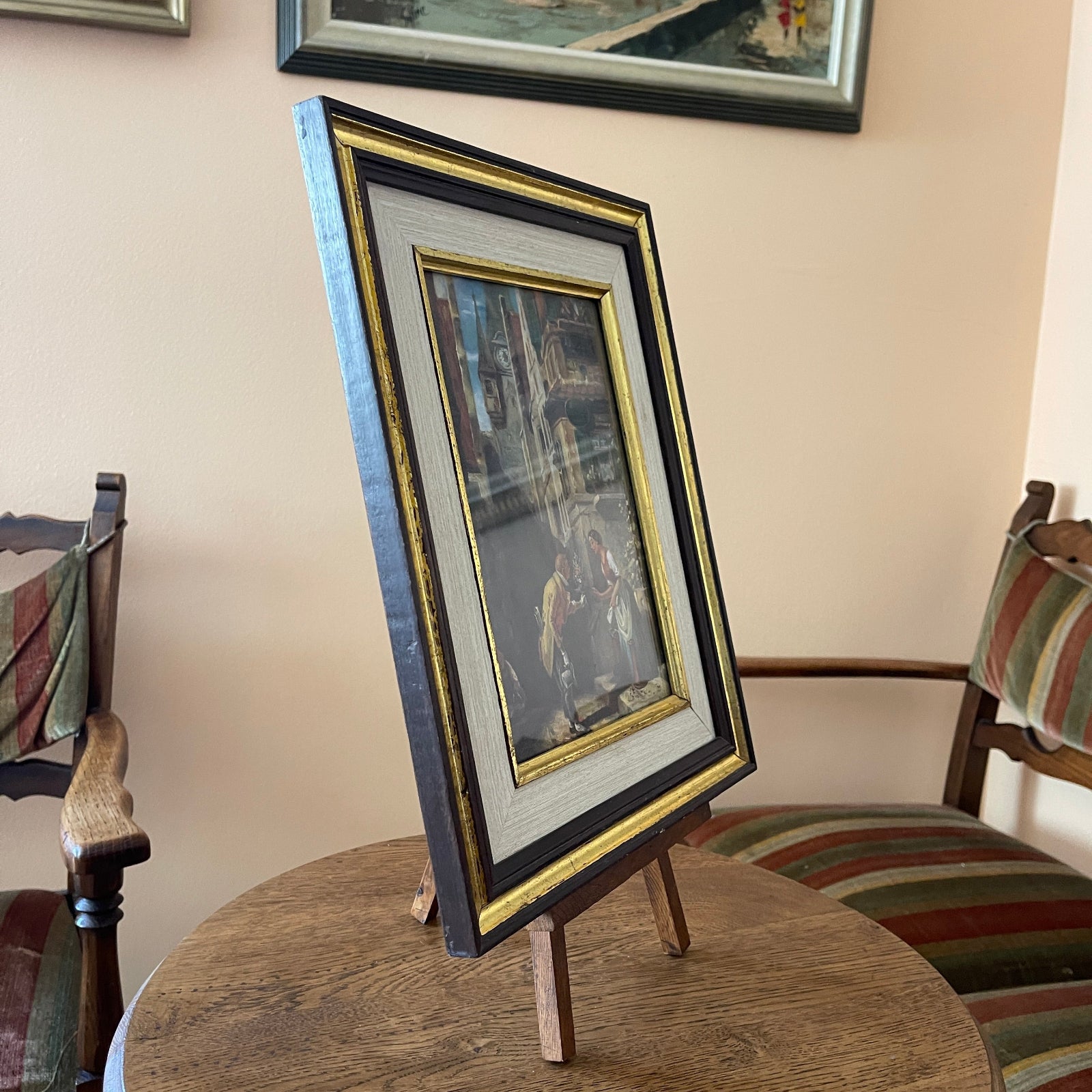 Framed picture on a stand with chairs and a wall with framed art in the background
