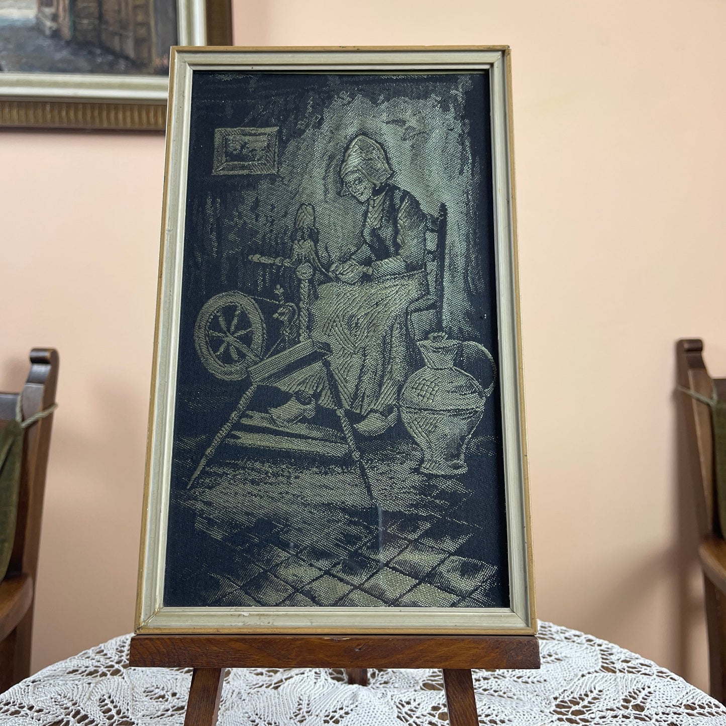 Framed artwork of a woman spinning yarn with a wheel on a lace tablecloth.