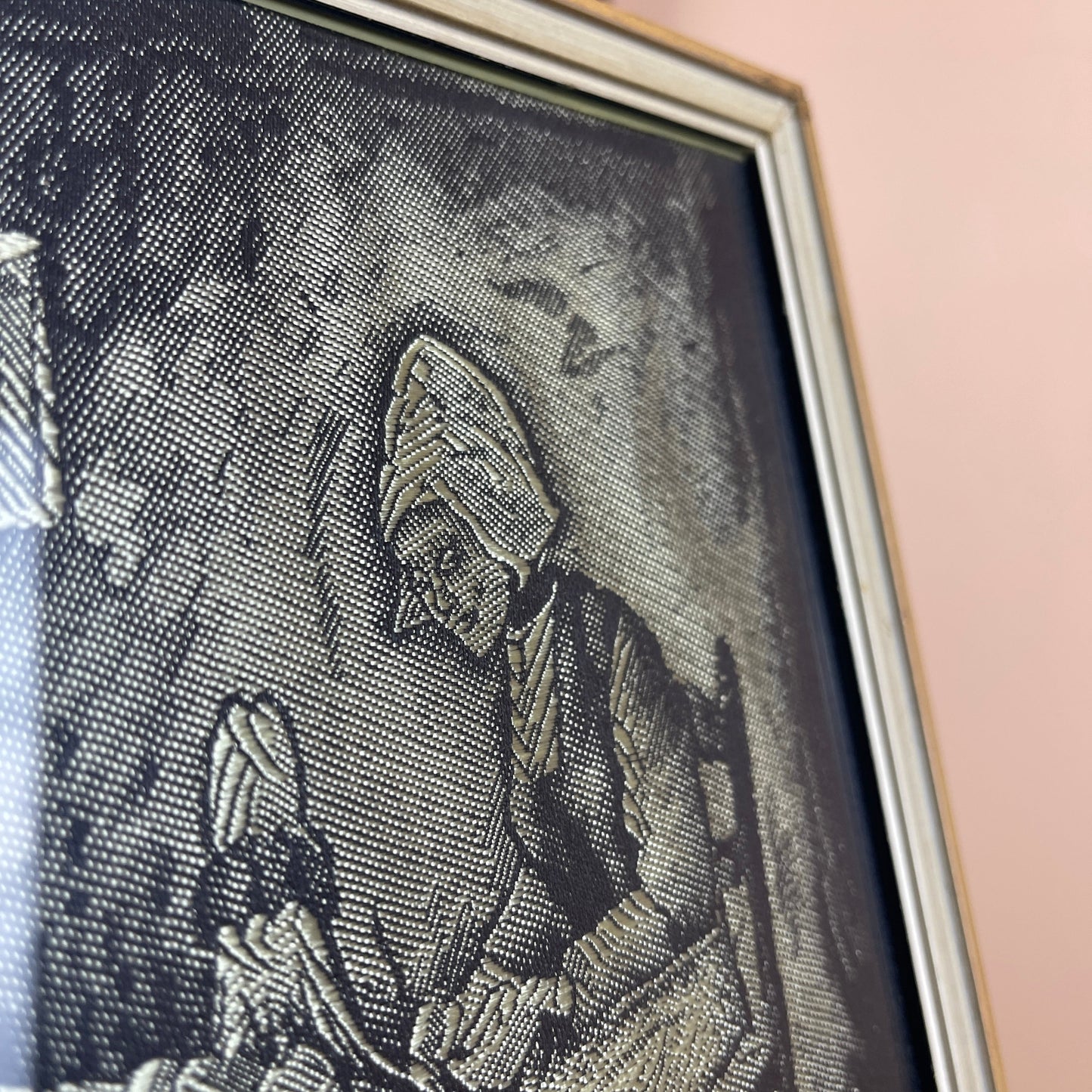 Framed black and white textile art of a person wearing a hat on a pink wall.