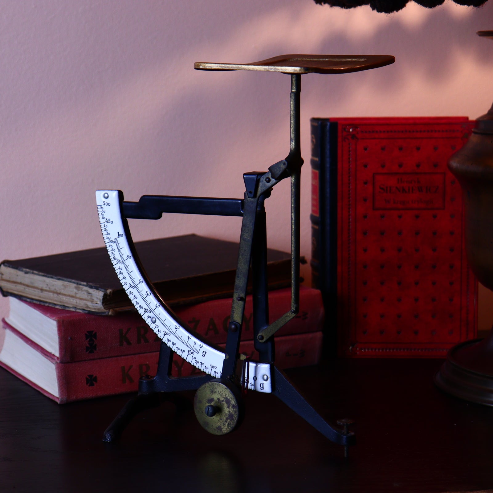 Vintage metal postal scale in black and gold colors, positioned on a flat surface with books and a lamp in the background.