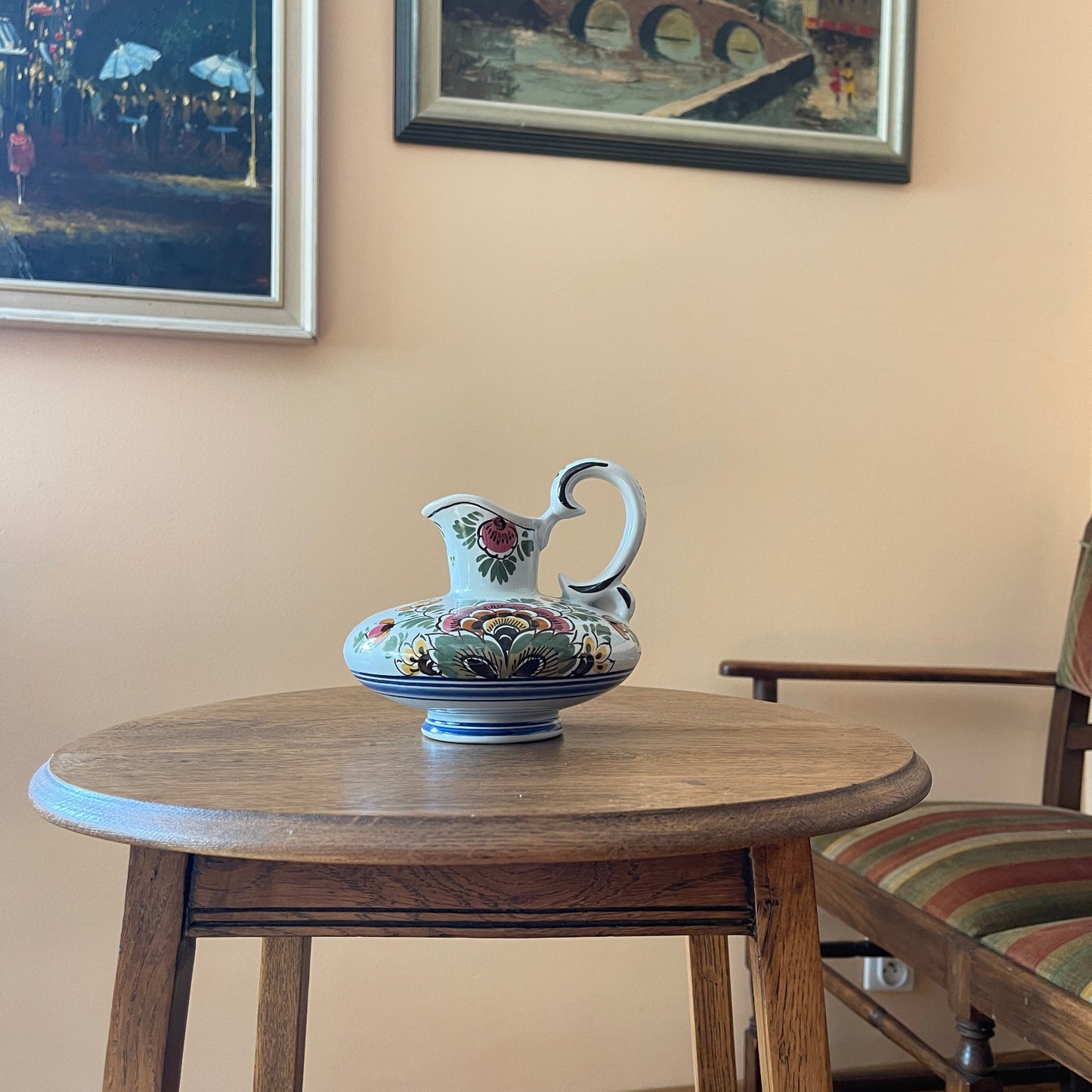 Decorative ceramic pitcher on a wooden table with framed artwork in the background.