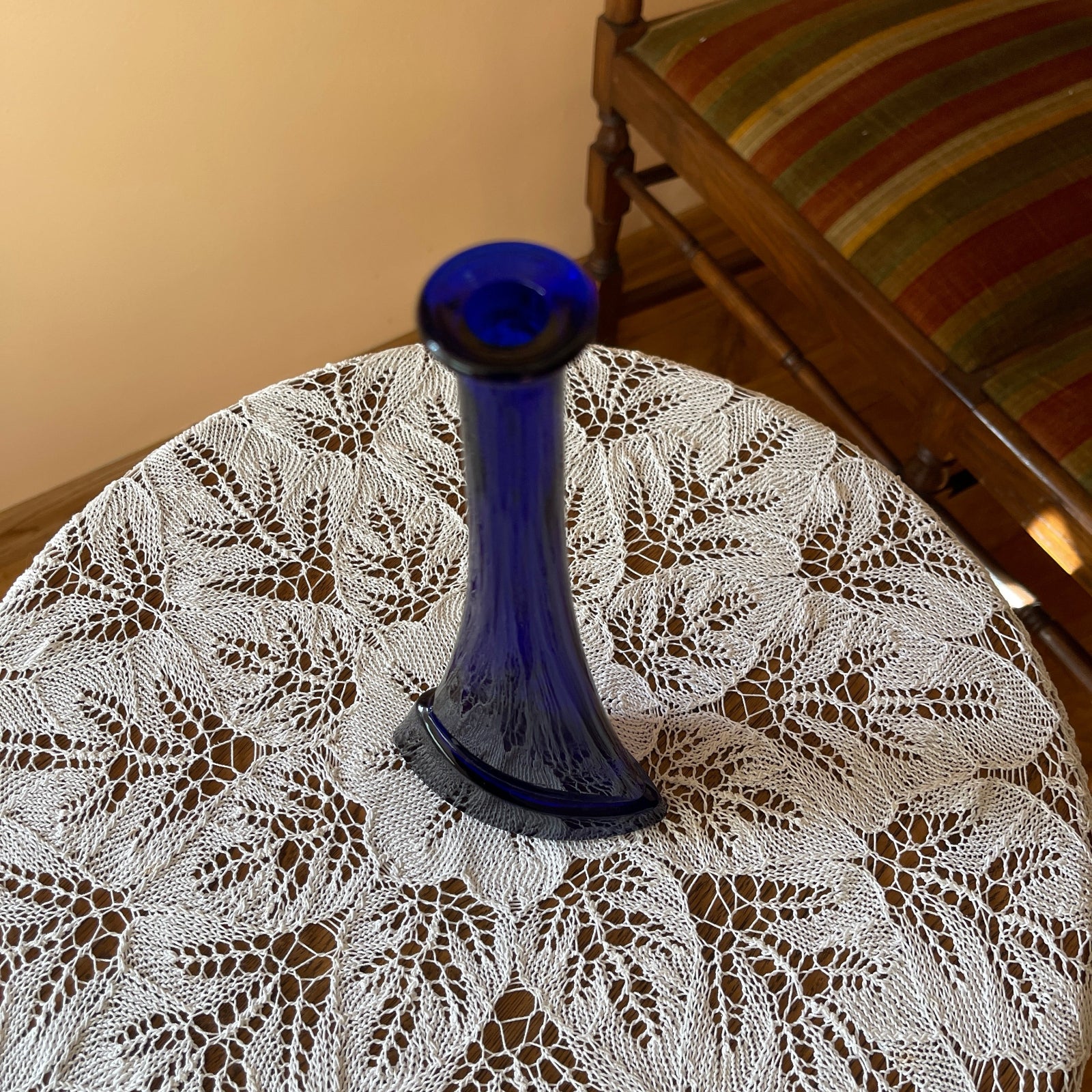 Blue glass vase on a lace tablecloth with a striped chair in the background