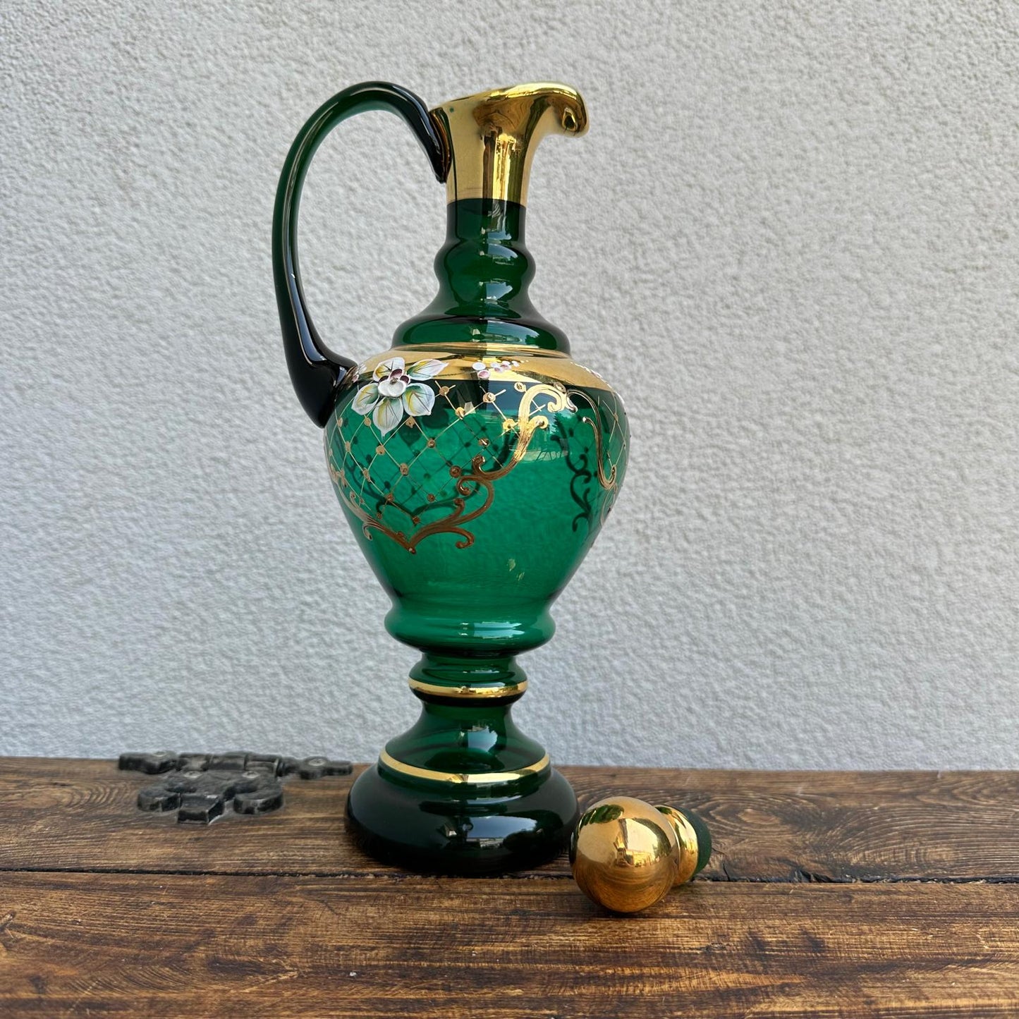 Decorative green pitcher with gold accents on a wooden surface against a light gray wall.