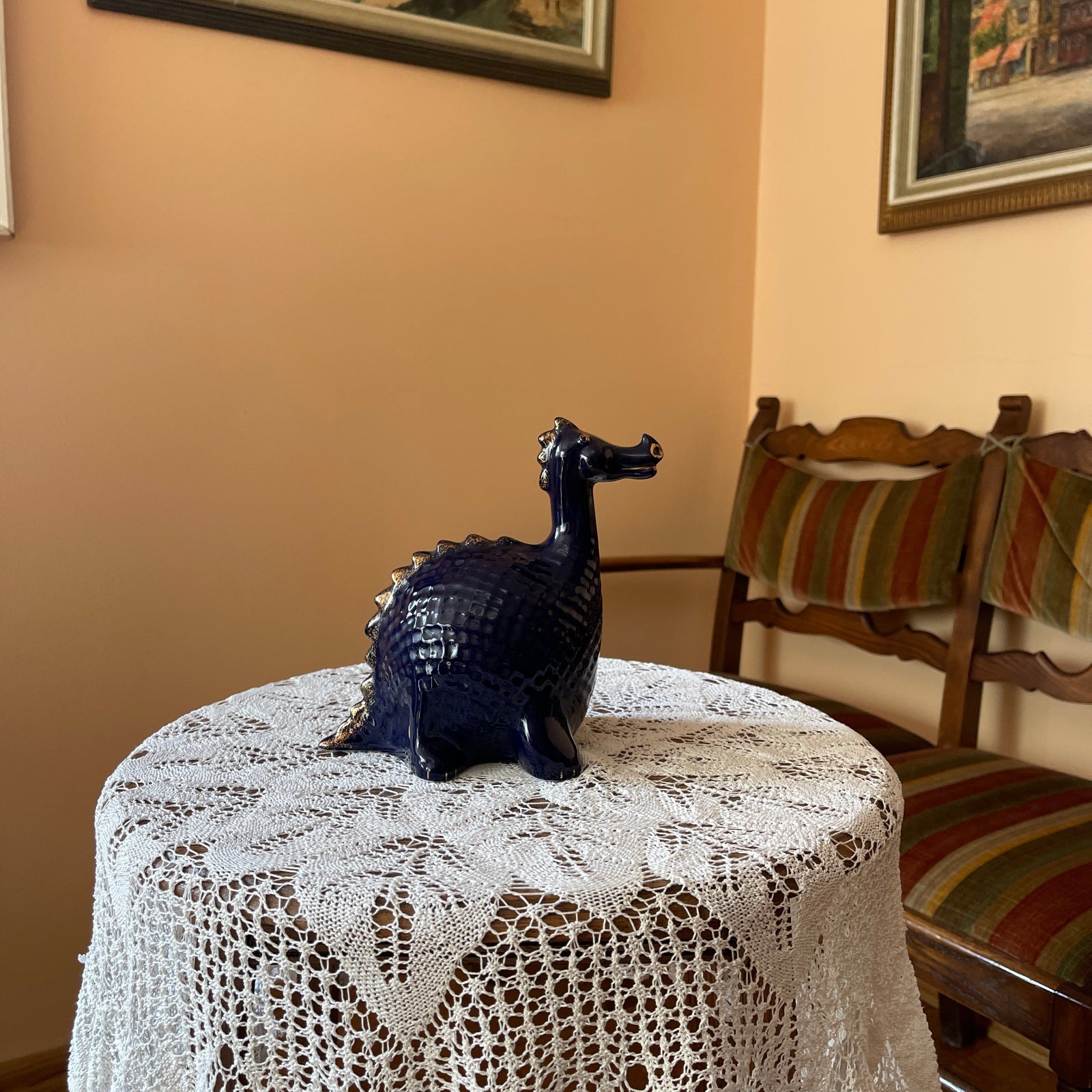 Decorative dragon figurine on a lace tablecloth with a chair and striped tablecloth in the background.