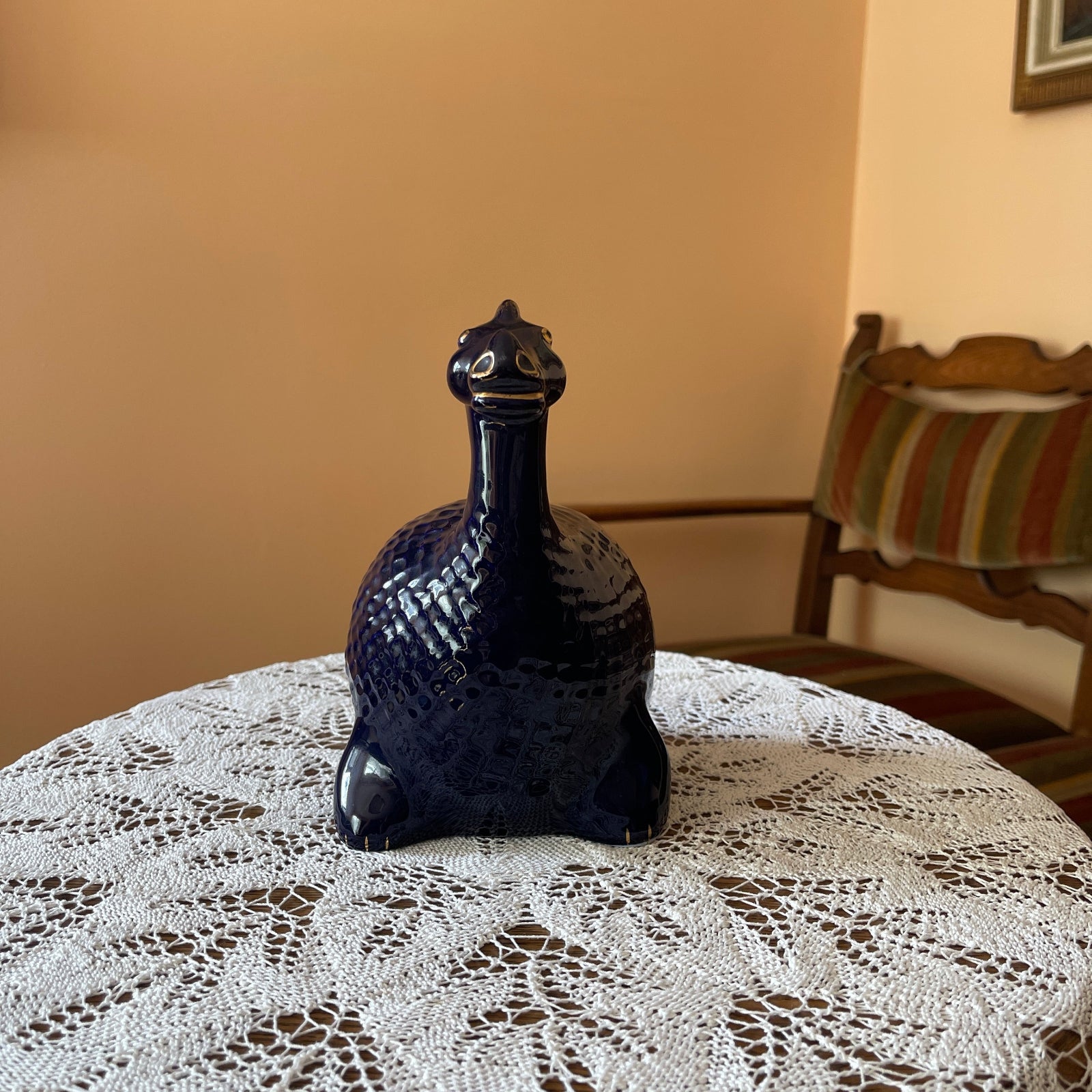 Black ceramic peacock figurine on a lace tablecloth with a chair and striped cushion in the background.