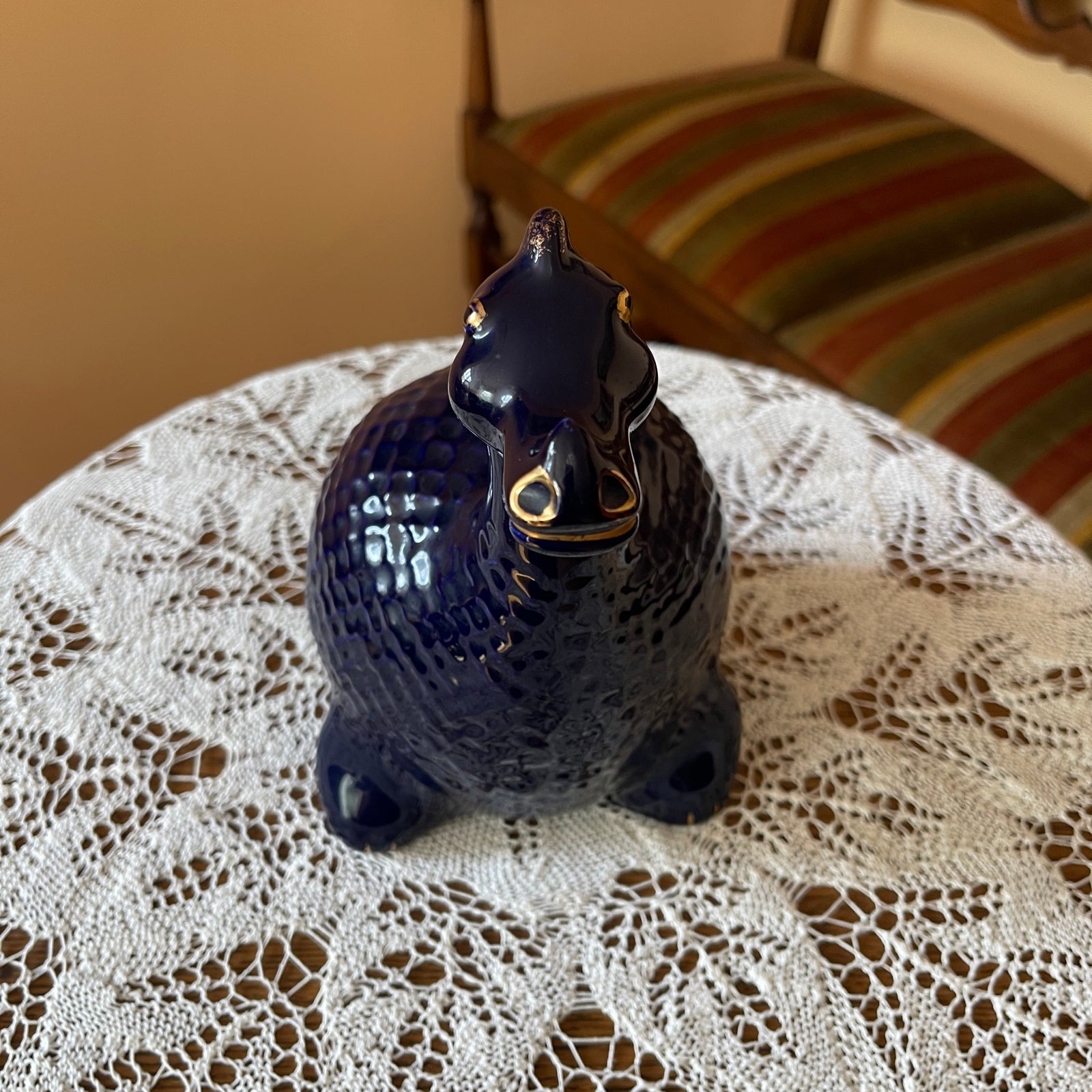Blue ceramic cat figurine on a lace tablecloth with a striped cushion in the background