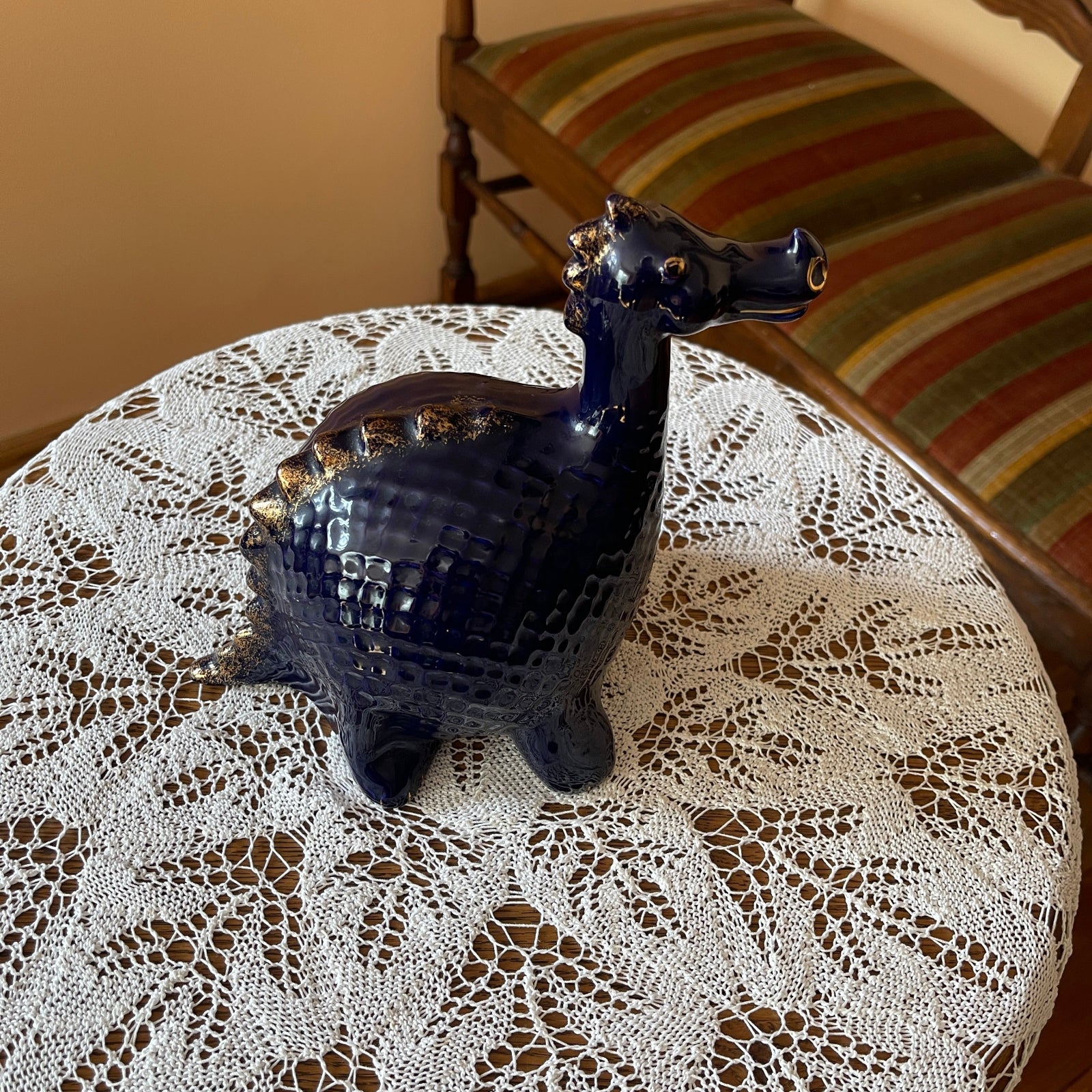 Blue ceramic peacock on a lace tablecloth with a striped chair in the background