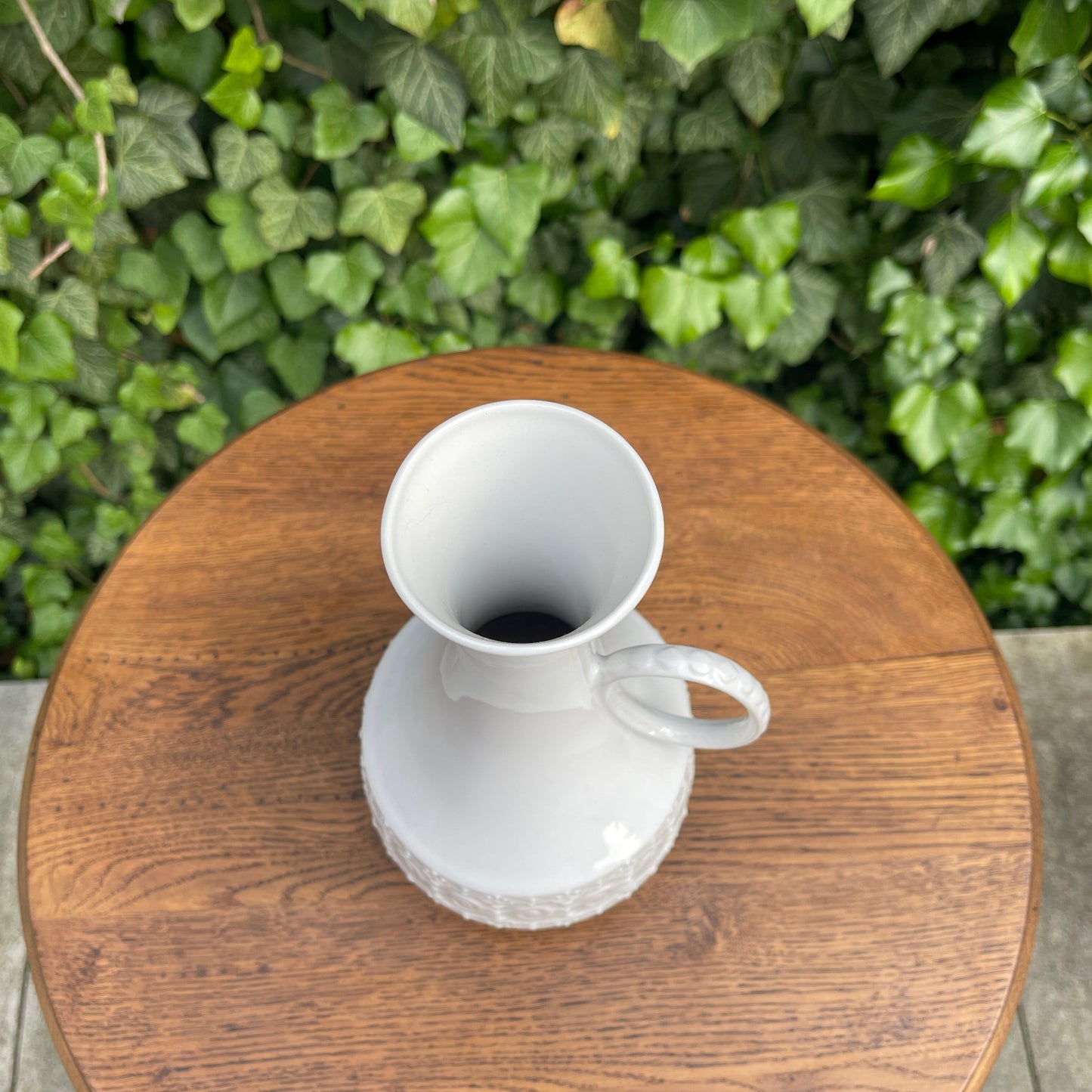 White ceramic vase on a wooden table with green foliage in the background