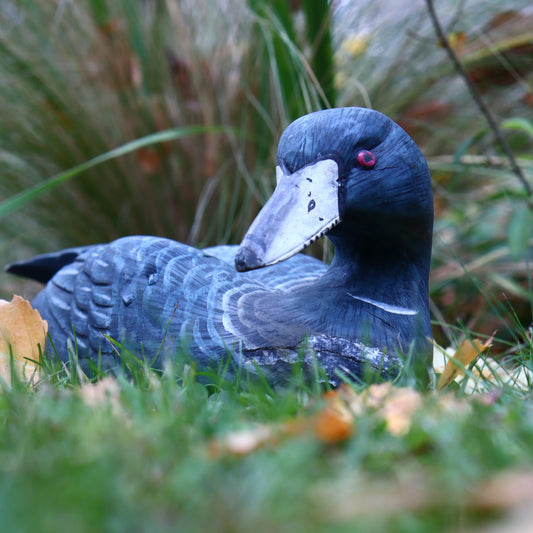 A vintage gray wooden figurine of a duck with a broken tip on one wing, resting on grass with leaves around.